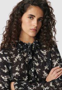 Blusa de gasa negra con estampado floral blanco, cuello fruncido y mangas largas, con puños elásticos y cierre con lazo.