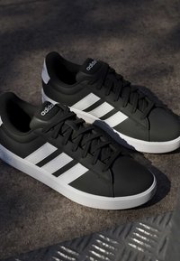 Paire de baskets Adidas noires avec des bandes et semelles blanches placées sur une surface grise texturée sous une lumière douce.