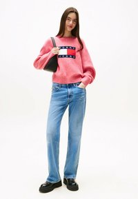 Rosa Strickpullover mit einem Marineblauen "TOMMY JEANS"-Logo, kombiniert mit hellblauen geraden Jeans und schwarzen Plateauschuhen.
