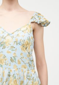 Αμάνικο ανοιχτό μπλε φόρεμα με κίτρινο λουλουδάτο σχέδιο και βολάν στους ώμους, φορεμένο στο πάνω μέρος του σώματος ενός ατόμου, με φόντο απλή επιφάνεια.