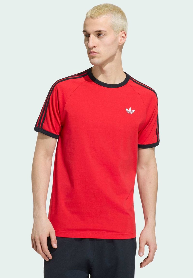 Young man with short blonde hair wearing a red Adidas t-shirt with black trim and stripes, looking to his left.