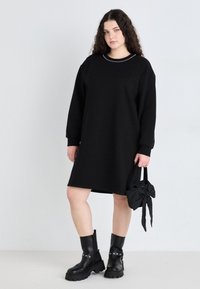 Čierne oversized šaty so sweatshiru s dlhými rukávmi, okrúhlym výstrihom a dekoračným lemovaním, skombinované s čiernymi masívnymi čižmami a malou čiernou taškou.