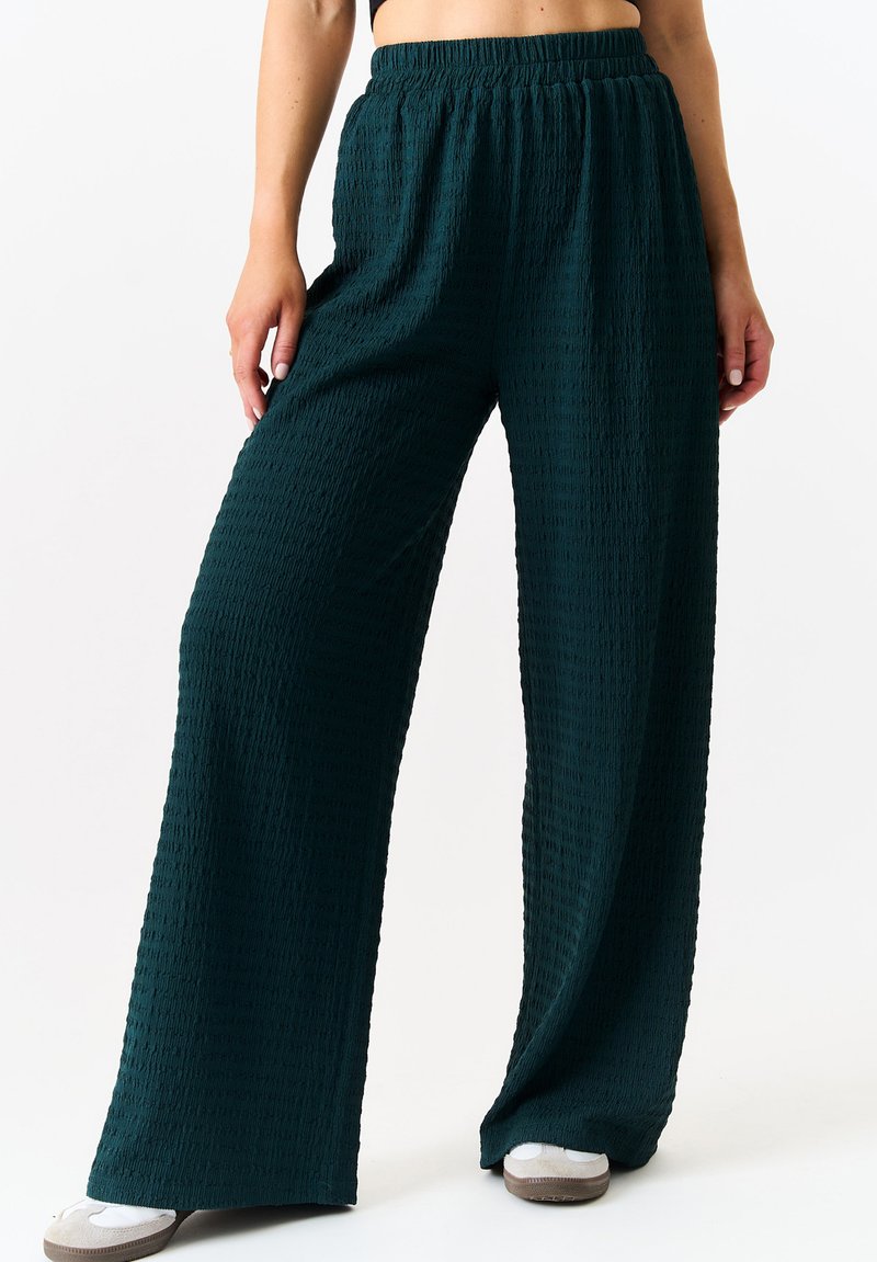 Wide-leg pants in dark teal with a textured fabric. Features an elastic waistband and a loose fit, paired with light-colored footwear.