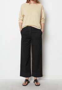 Beige svetr s dlouhým rukávem a černé široké kalhoty z hladkého materiálu. Kalhoty mají boční kapsy a uvolněný střih; sandály jsou jednoduché černé.