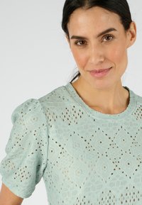 Haut vert menthe clair à manches courtes en dentelle texturée avec un motif en losange et un col rond ; les manches bouffantes ajoutent du volume.
