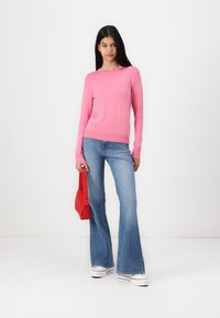 Roza pletena jopica z dolgimi rokavi, okroglim ovratnikom, v kombinaciji z razširjenimi modrimi kavbojkami. Drži rdečo torbico in stoji na beli podlagi.