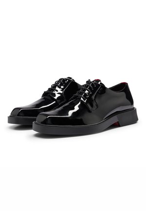 Paire de chaussures habillées noires en cuir brillant, à lacets, avec des bouts ronds lisses et des petits talons en caoutchouc noir sur fond blanc.