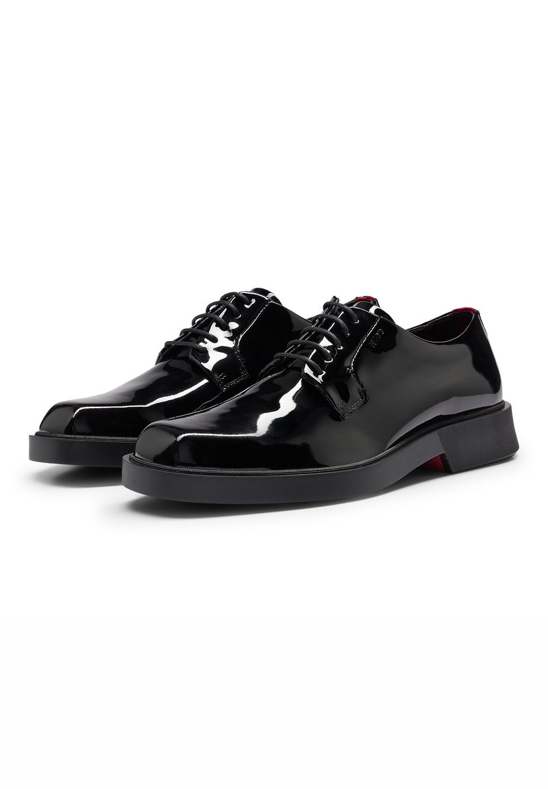 Paire de chaussures habillées noires en cuir brillant, à lacets, avec des bouts ronds lisses et des petits talons en caoutchouc noir sur fond blanc.