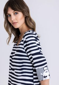 Vrouw met schouderlang bruin haar, gekleed in een zwart-wit gestreept shirt met een grafisch gezichtsontwerp op de mouw, tegen een witte achtergrond.