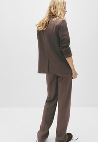 Žena s dugom plavom kosom nosi široko smeđi odijelo s podvrnutim rukavima i širokim hlačama, stojeći nasuprot bijeloj pozadini.