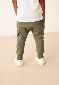 Maslinasto zelene trenirka hlače s rebrastim manžetama i bočnim džepovima, uparene s bijelim tenisicama, pogled s leđa. Glatka pamučna tekstura.