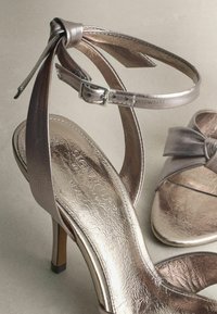 Metallic high-heeled sandals feature a textured strap, ankle buckle, and a bow detail. The color is a subtle silver with a glossy finish.
