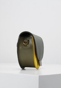 Mała usztywniana torebka z oliwkowozielonymi bokami, szarą klapką i żółtym wnętrzem, stojąca pionowo na białej powierzchni.