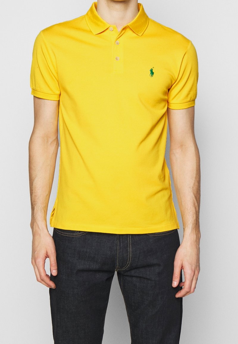 Homme portant un polo jaune vif à manches courtes avec un logo vert sur la poitrine et un jean en denim foncé, debout devant un fond clair.