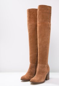 Alte stivali in suede marrone con tacco conico, cerniere laterali e punte arrotondate appoggiate su uno sfondo chiaro e semplice.