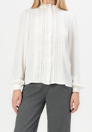 Woman wearing a long-sleeve white blouse with lace and pleat details, paired with dark grey tailored pants, shown from neck to mid-thigh.
