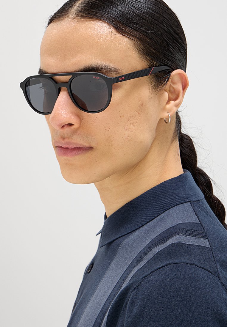Schwarze rechteckige Sonnenbrille mit dunklen Gläsern und einem strukturierten schwarzen Arm mit roten Akzenten. Dazu getragen wird ein navyblaues Poloshirt.