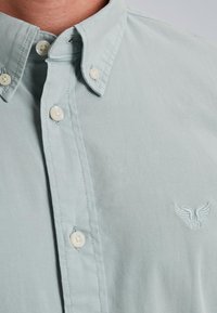 Camisa de botones azul claro de algodón con cuello, que presenta botones blancos y un logotipo bordado en el pecho. Textura suave.