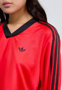 Raudona sportinė maikė su juodais akcentais, V formos apykaklė ir vertikalios juostos. Pagrindinis juodas Adidas logotipas ant krūtinės.