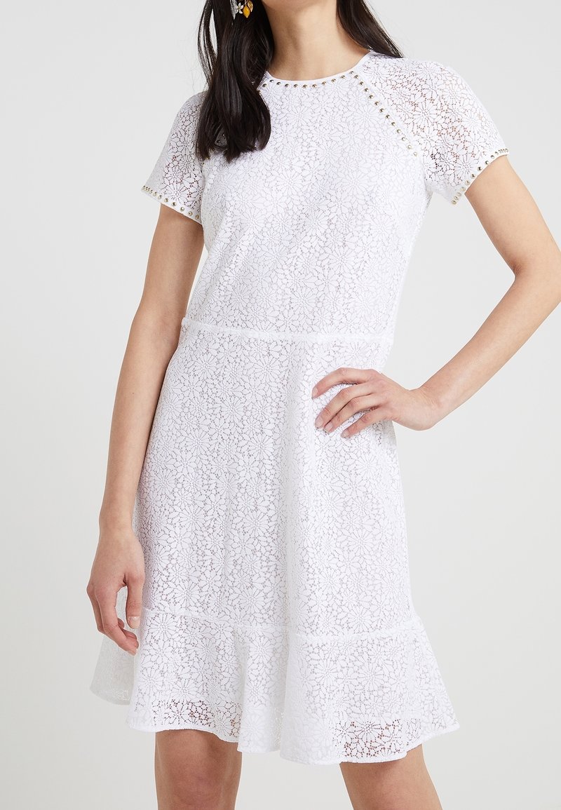 Femme portant une robe en dentelle blanche à manches courtes avec motif floral et détails cloutés sur les manches et le décolleté, main sur la hanche.