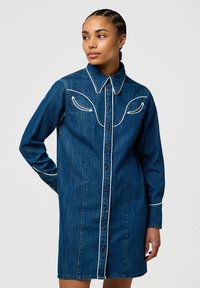 Abito camicia in denim caratterizzato da un colletto classico, chiusura frontale con bottoni, cuciture bianche di contrasto sul giogo e sui polsini, e una silhouette aderente.