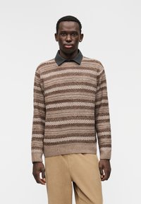 Hnědý a béžový pruhovaný pletený svetr s tmavým límečkem, v kombinaci s béžovými manšestrovými kalhotami. Má žebrované manžety a lem.