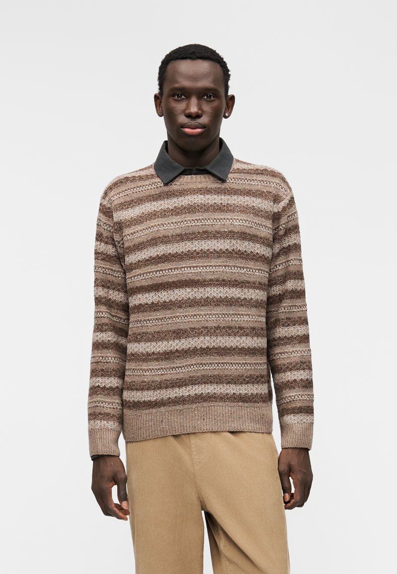 Hnědý a béžový pruhovaný pletený svetr s tmavým límečkem, v kombinaci s béžovými manšestrovými kalhotami. Má žebrované manžety a lem.