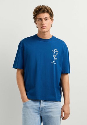 Junger Mann mit lockigem Haar, der ein blaues T-Shirt mit dem Schriftzug „THE LADS“ und einer Grafik eines Tennisspielers trägt, kombiniert mit hellblauen Jeans.