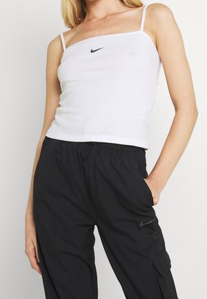 Vrouw draagt een wit Nike spaghetti bandjes tanktop en zwarte Nike cargobroek, met haar hand in de zak, staande tegen een lichte achtergrond.