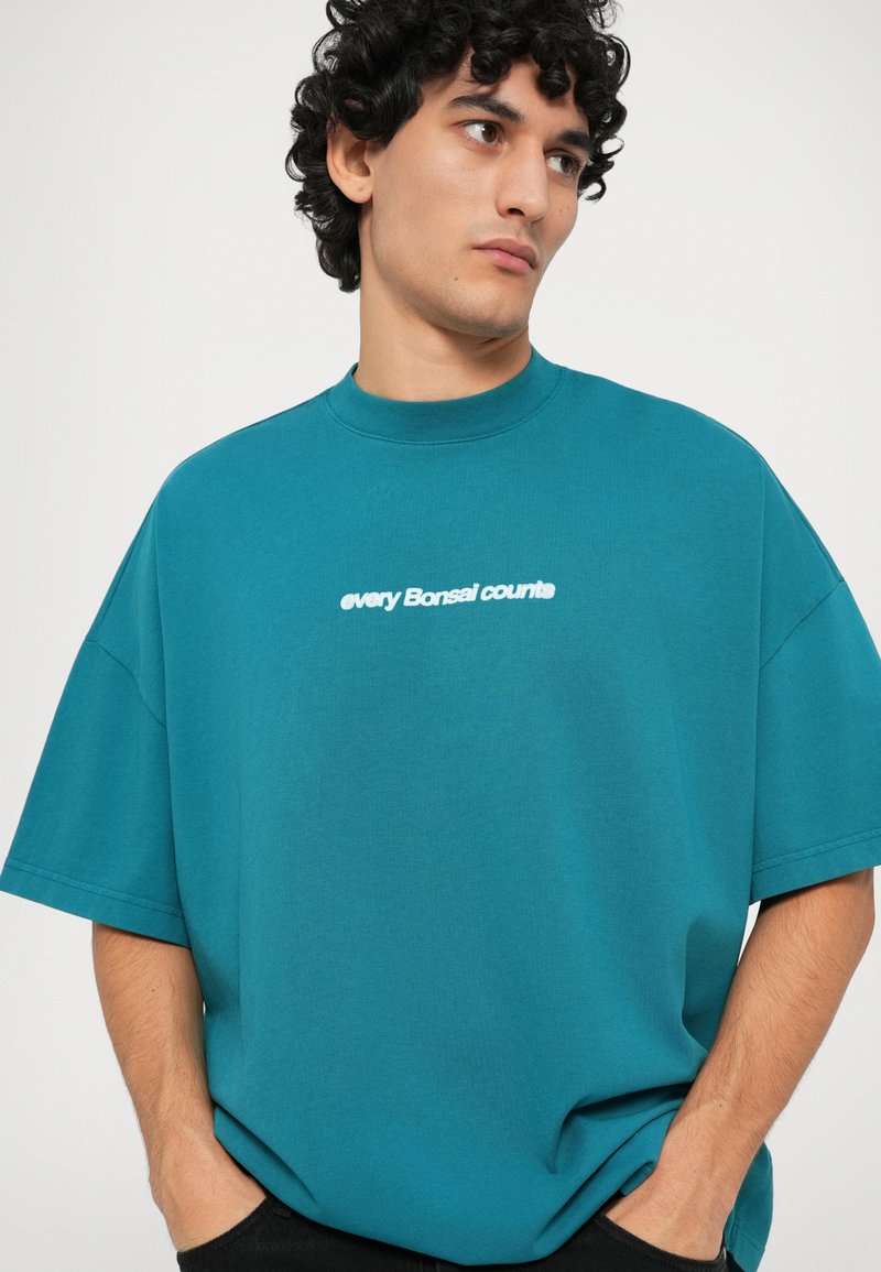 T-shirt surdimensionné teal en mélange de coton avec un col montant et des manches courtes. Il présente du texte blanc "chaque Bonsai compte" sur le devant.