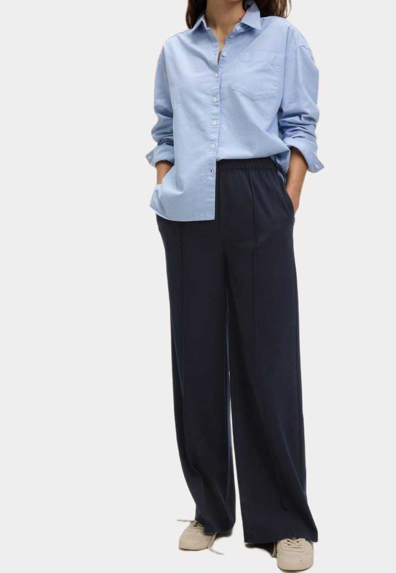 Chemise à boutons bleu clair avec une poche, associée à un pantalon bleu marine taille haute et jambes larges, ainsi que des chaussures de couleur claire. Textures de tissu lisses.
