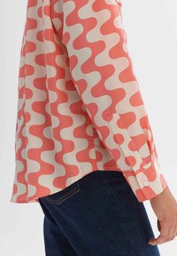 Long sleeve shirt with coral and cream abstract wave pattern worn with dark blue jeans.