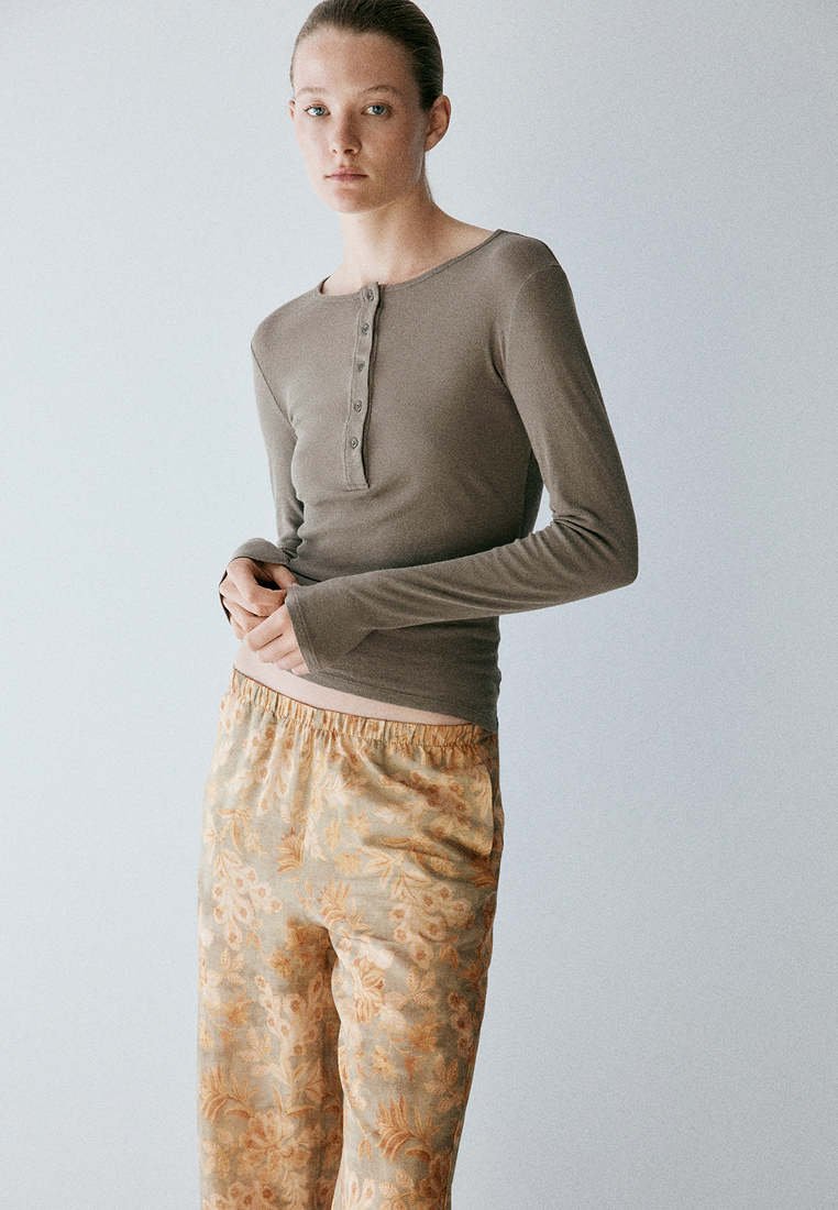 Jeune femme portant un haut taupe à manches longues ajusté et un pantalon beige ample à motif floral, debout devant un fond clair uni.