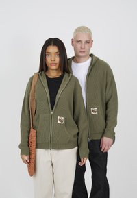 Olive green ribbed hoodies with front pockets and logo patches, worn over black and white tops. Light brown crocheted bag.