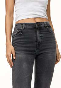 Kvinde iført sorte højtaljede skinny jeans og en opsmøget hvid ærmeløs top, med hænderne hvilende ved hendes sider.