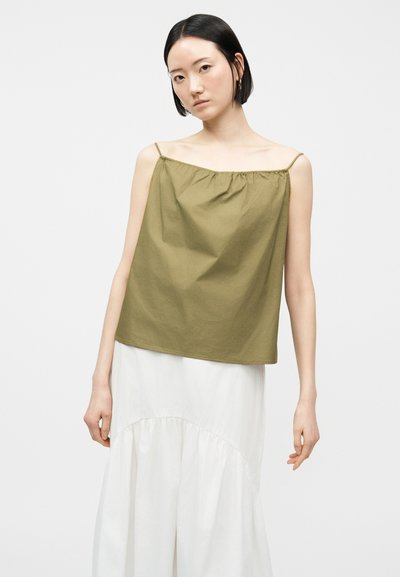 Νεαρή γυναίκα με κοντά μαύρα μαλλιά, που φοράει αμάνικο τοπ ελιάς και μακριά λευκή φούστα, μπροστά σε απλό φωτεινό φόντο.