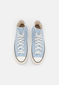 Vysoké tenisky světle modré plátno s bílými kulatými tkaničkami, bílým gumovým ochranným krytem špičky a černým pruhovaným detailem na podrážce.