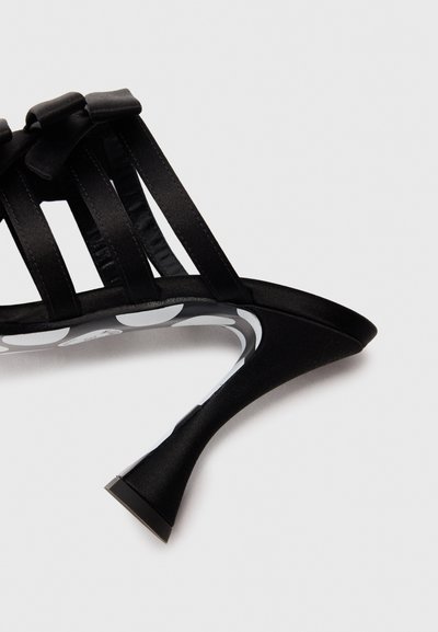 Escarpin noir à talons hauts avec trois brides et de petits nœuds, présentant un talon courbé et une semelle intérieure à pois, sur un fond blanc uni.