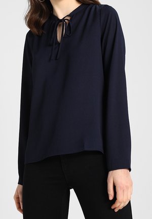 Femme portant un chemisier bleu marine à manches longues avec un petit détail de lien à l'avant au niveau du col, associé à un pantalon noir.