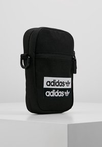 Černá plátěná crossbody taška s dvěma bílými nášivkami loga Adidas, obdélníkový tvar, zipové zapínání a malá rukojeť na boku.