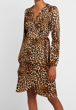 Femme portant une robe portefeuille à manches longues imprimée léopard, col en V et longueur genou, debout sur un fond uni.