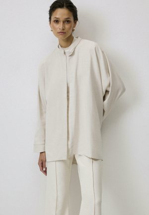 Blusa oversized beige claro con cuello alto, hombros caídos y mangas anchas, hecha de una suave tela texturizada. Diseño minimalista.