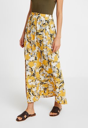 Gul maxi nederdel med hvidt og sort blomsterprint, båret med olivengrøn top og mørkebrune vævede sandaler.