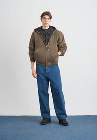 Smeđa dukserica na patentni zatvarač s crnim podstavom, podesivim kapuljačom i prednjim džepovima; uparena s plavim širokim trapericama i crnim cipelama.