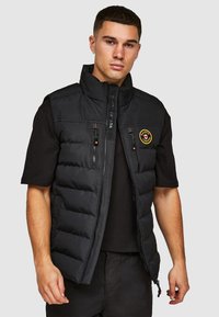Zwarte puffer gilet met een hoge kraag, voorzijde met rits en twee ritszakken. Bevat een cirkelvormig logo op de linkerkant van de borst. Soepele textuur.