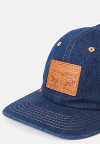 Ciemnoniebieska czapka z dżinsu z brązową skórzaną naszywką z wytłoczonym tekstem i grafiką. Widoczne detale szycia na daszku.