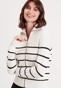 Pull en tricot blanc zippé avec des rayures horizontales noires, texture côtelée, col montant et manches longues avec les trous pour les pouces.
