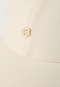 Kremowa czapka z daszkiem z teksturowanego materiału, z ozdobnym złoto-białym logo na przodzie. Strukturalny kształt z zakrzywionym daszkiem.
