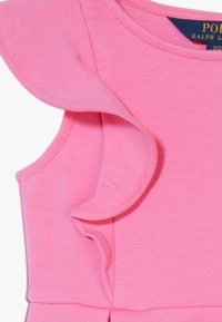 Rosa ärmlös klänning med volangdetaljer på axlarna, gjord av mjukt bomullsmaterial; har en rund halsringning och stickade detaljer.