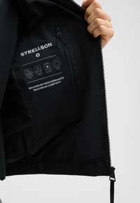 Schwarz gefütterte Jacke aus wasserdichtem Stoff, mit Reißverschlusstasche und bedruckten Pflegeikonen. Weiche Textur mit perforierten Details entlang des Futters.